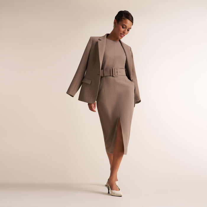 A woman models a taupe-colored two-piece set consisting of a belted midi dress and a matching blazer, paired with white pointed-toe heels against a solid light beige studio background.