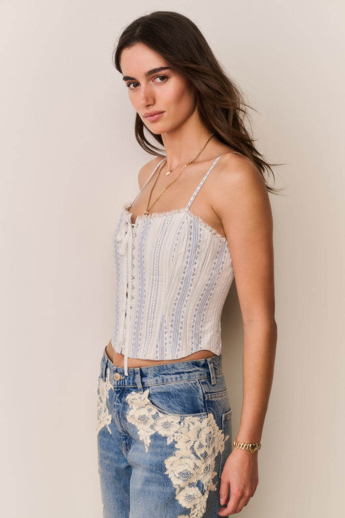 Woman wearing a white and blue stripe lace-up top with thin straps and denim pants with lace details against a beige background