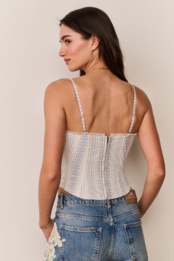 Woman wearing a white and blue stripe lace-up top with thin straps and denim pants with lace details against a beige background