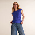 A woman modeling a sleeveless, one-shoulder royal blue top with gathered detailing, paired with blue jeans, against a plain light beige studio background. | Lead