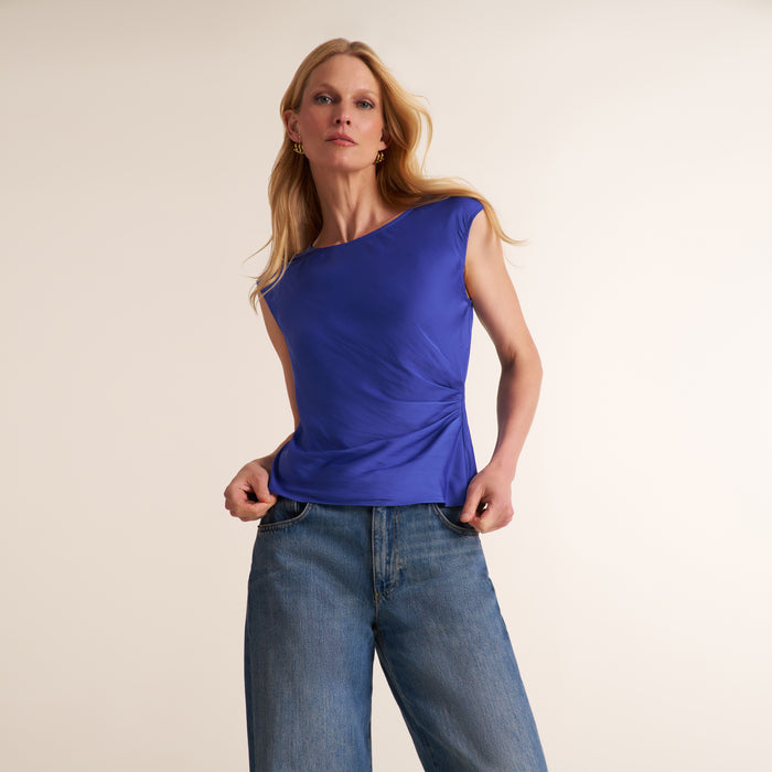 A woman modeling a sleeveless, one-shoulder royal blue top with gathered detailing, paired with blue jeans, against a plain light beige studio background. | Lead