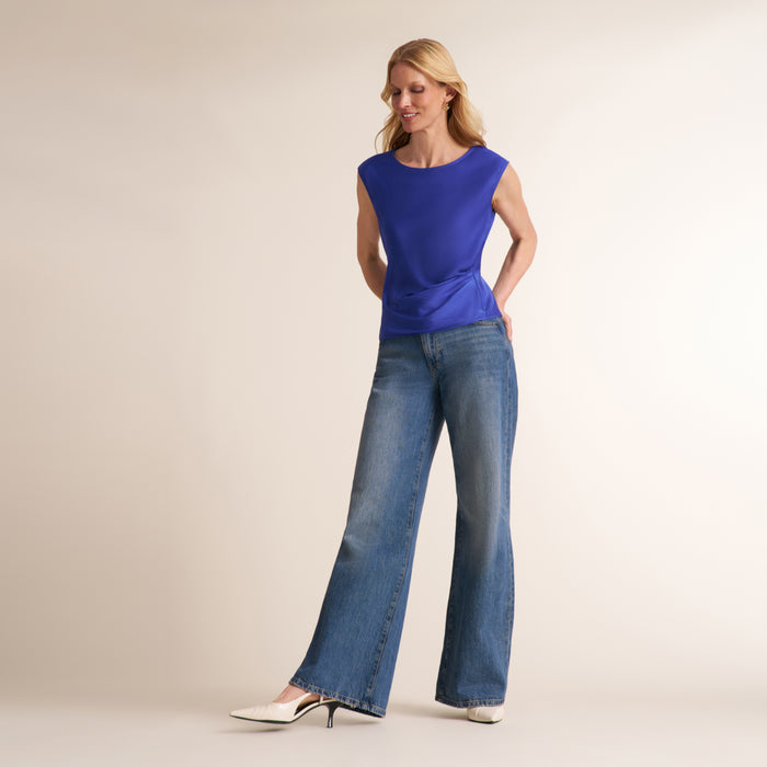 A woman modeling a sleeveless royal blue top and wide-leg blue jeans with white pointed-toe heels, standing against a plain beige studio background.