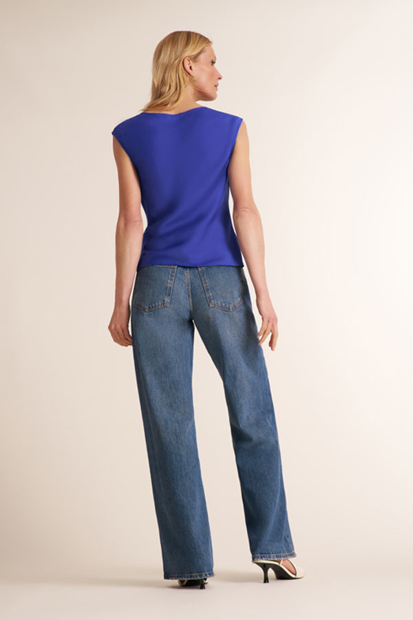 A woman shown from the back wearing a sleeveless royal blue top and blue jeans, standing against a plain light beige studio background. | Exclude
