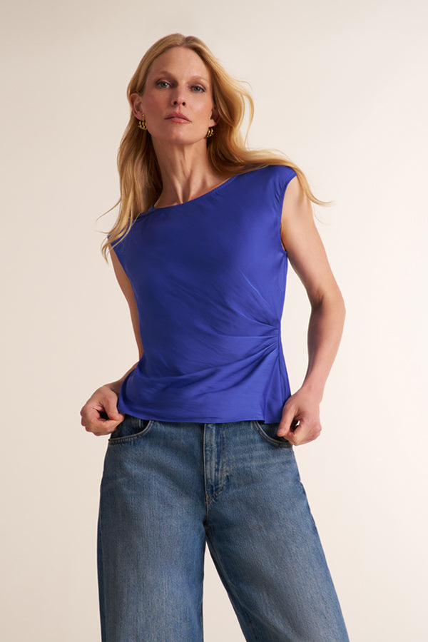 A woman modeling a sleeveless, one-shoulder royal blue top with gathered detailing at the waist, paired with blue jeans, against a plain beige studio background. | Exclude | Grid