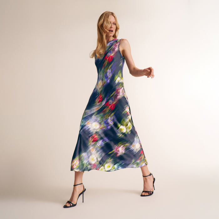 A woman modeling a sleeveless midi dress with a dark blue base and a colorful abstract floral print, paired with black strappy high-heeled sandals against a plain beige studio background. | Lead