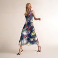 A woman modeling a sleeveless midi dress with a dark blue base and a colorful abstract floral print, paired with black strappy high-heeled sandals against a plain beige studio background. | Lead