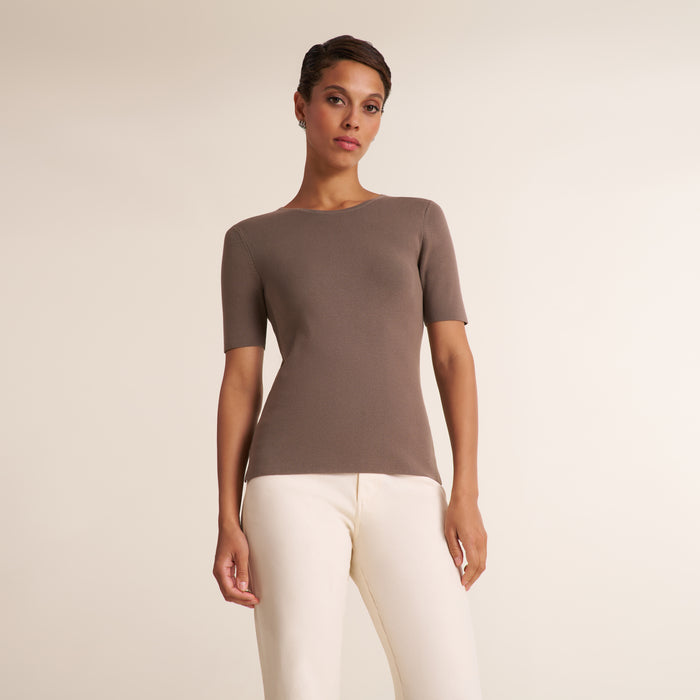 A woman modeling a short-sleeve ribbed knit top in taupe brown with off-white pants, standing against a plain light beige studio background. | Lead