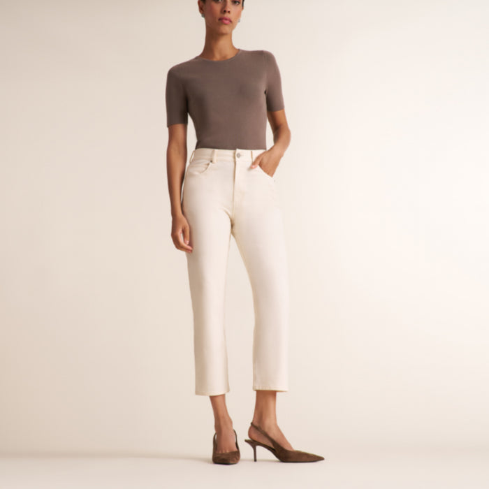 A model wearing a fitted taupe short-sleeve top, off-white cropped trousers, and brown pointed-toe heels, standing against a plain warm beige studio background.