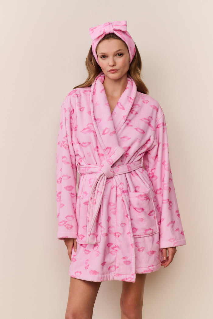 Woman wearing a pink robe with kiss print pattern and matching headband against a beige background