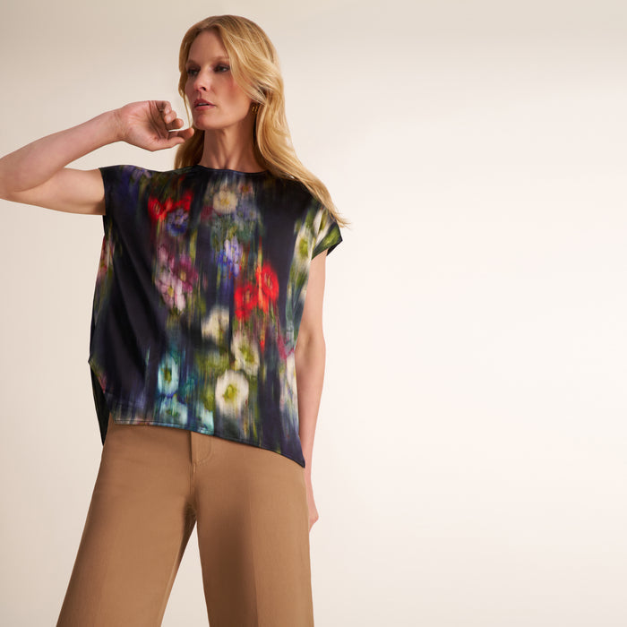 A woman modeling a short-sleeve top with a dark navy base and a colorful floral print, paired with tan trousers, against a plain light beige studio background. | Lead