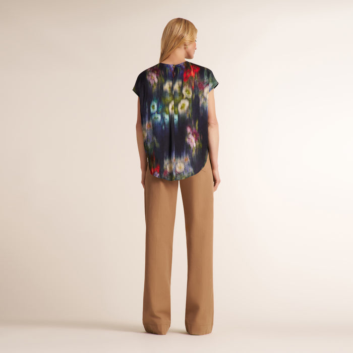 A woman stands with her back to the camera wearing a short-sleeve top with a dark abstract floral print and tan wide-leg pants against a plain beige studio background.
