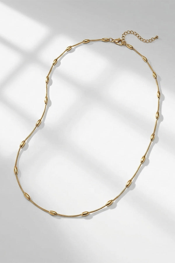 A gold chain necklace with small, evenly spaced teardrop-shaped links, displayed on a light gray surface with soft window-box shadows. | Still | Exclude