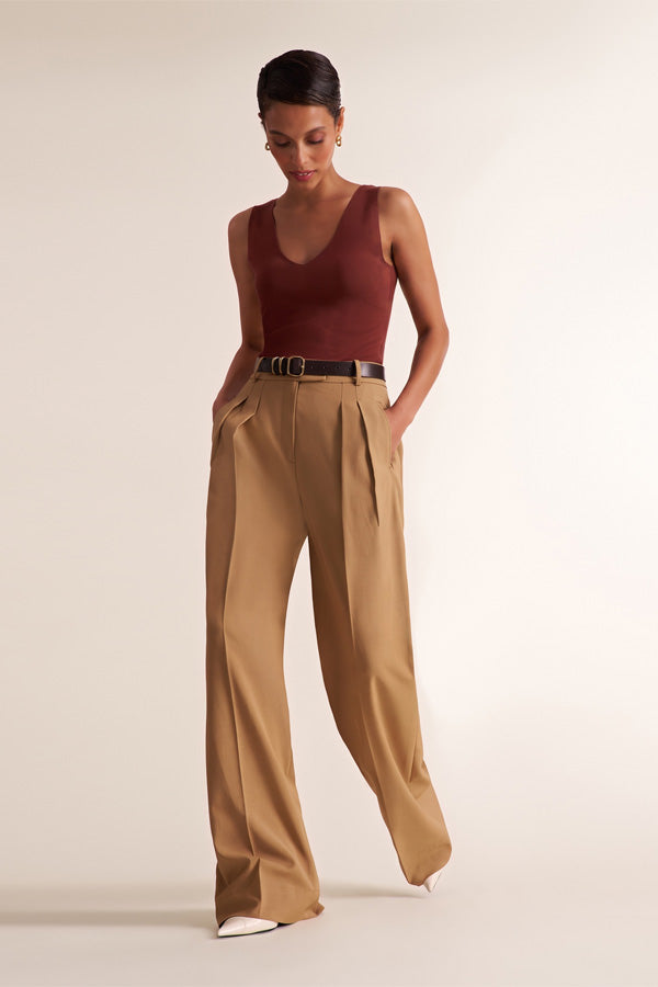 A model wearing a sleeveless burgundy top and high-waisted tan wide-leg trousers with a thin black belt, standing against a plain light beige studio background. | Exclude