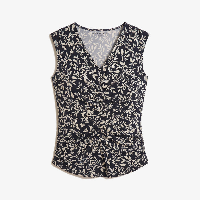 A sleeveless wrap-style top with an allover beige leaf and floral pattern on a dark navy background, featuring a V-neckline and gathered waist details, shown against a light gray background.