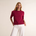 A woman modeling a short-sleeve crewneck sweater in deep magenta, paired with white trousers, against a plain light beige studio background. | Lead
