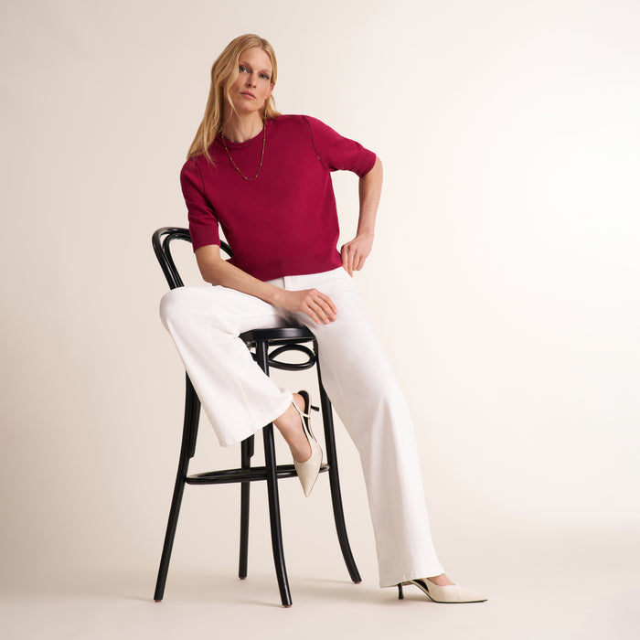 A woman sits on a black bar stool wearing a short-sleeve burgundy knit top and white wide-leg trousers, photographed against a plain warm off-white studio background.