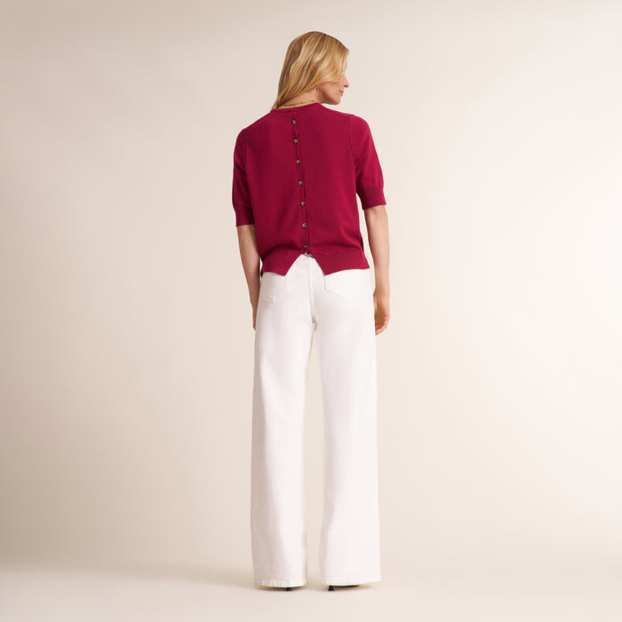 A woman viewed from the back wearing a short-sleeve burgundy knit top with a buttoned back and white wide-leg trousers, standing against a plain warm beige studio background.