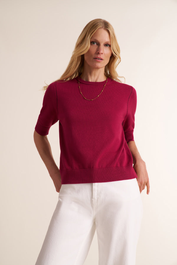 A woman modeling a short-sleeve crewneck sweater in deep magenta, paired with white trousers, against a plain light beige studio background. | Exclude | Grid