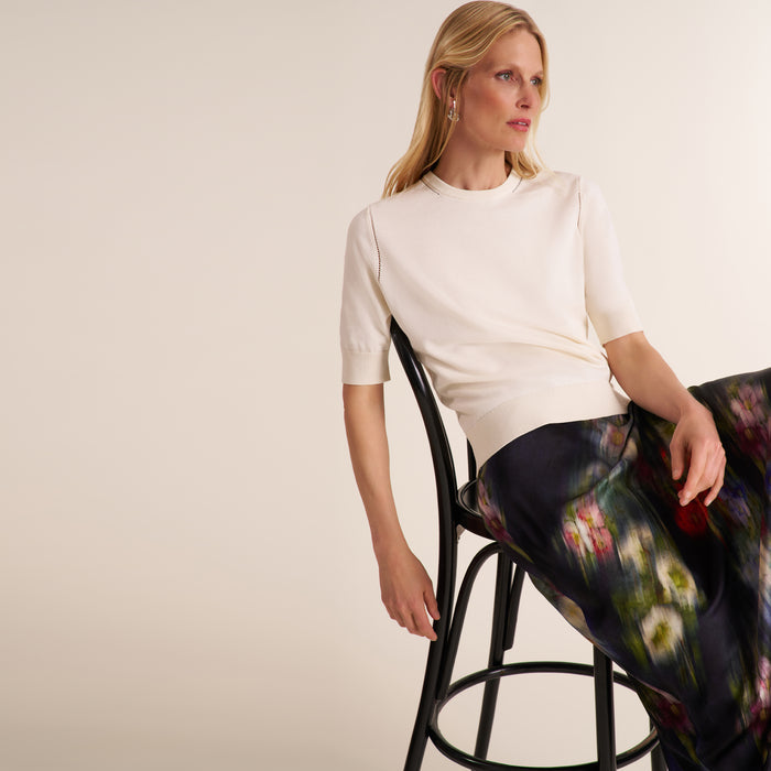 A woman seated on a black metal stool wearing a fitted short-sleeve cream top and a dark floral skirt, photographed against a plain warm off-white studio background. | Lead
