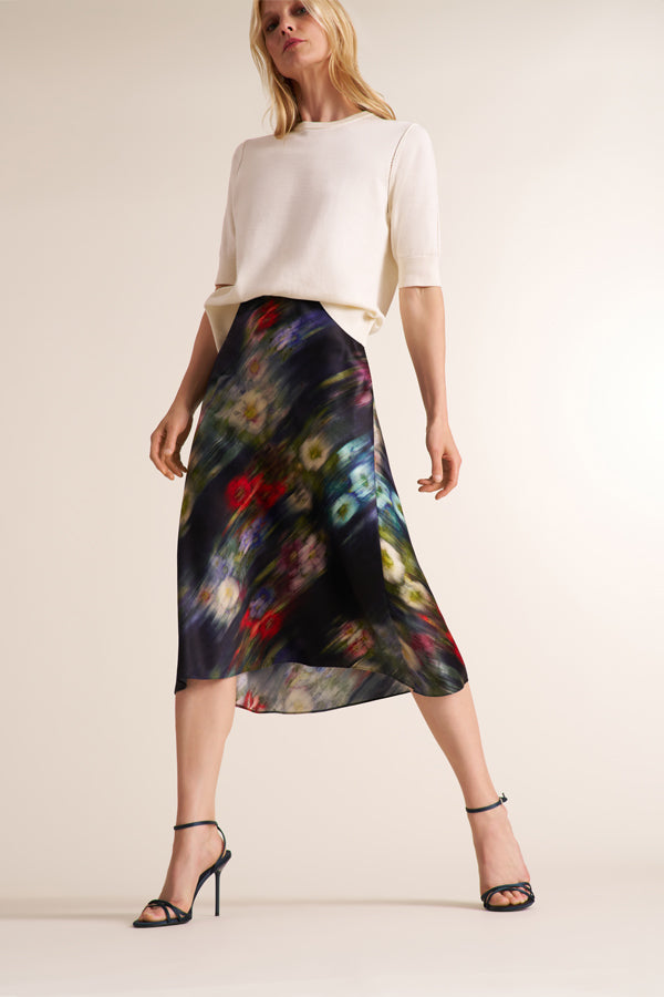 A model wearing a cream short-sleeve knit top and a black midi skirt with a vibrant, multicolored abstract floral print, paired with black strappy high-heeled sandals against a solid light beige background. | Exclude