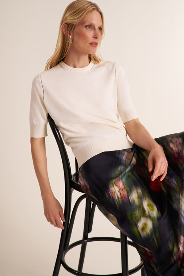 A woman seated on a black metal stool wearing a cream short-sleeve knit top and a dark floral skirt, photographed against a plain light beige studio background. | Exclude | Grid