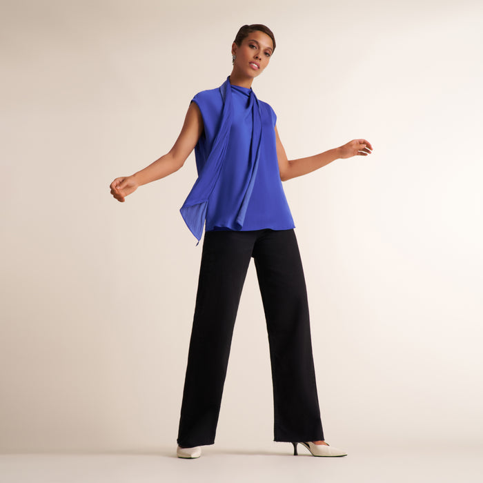 A model wearing a sleeveless, high-neck royal blue draped blouse and black wide-leg trousers, standing against a solid light beige studio background.