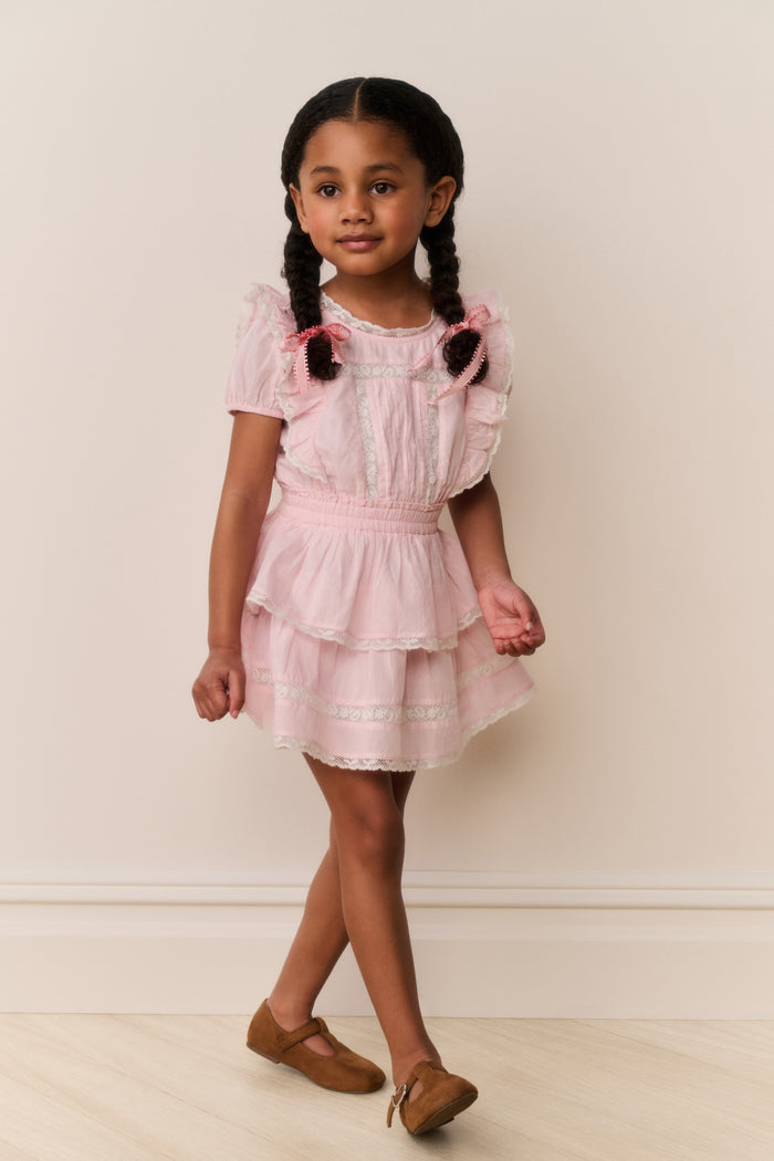 Young girl wearing a pink dress with ruffled sleeves and layers, standing against a plain background.