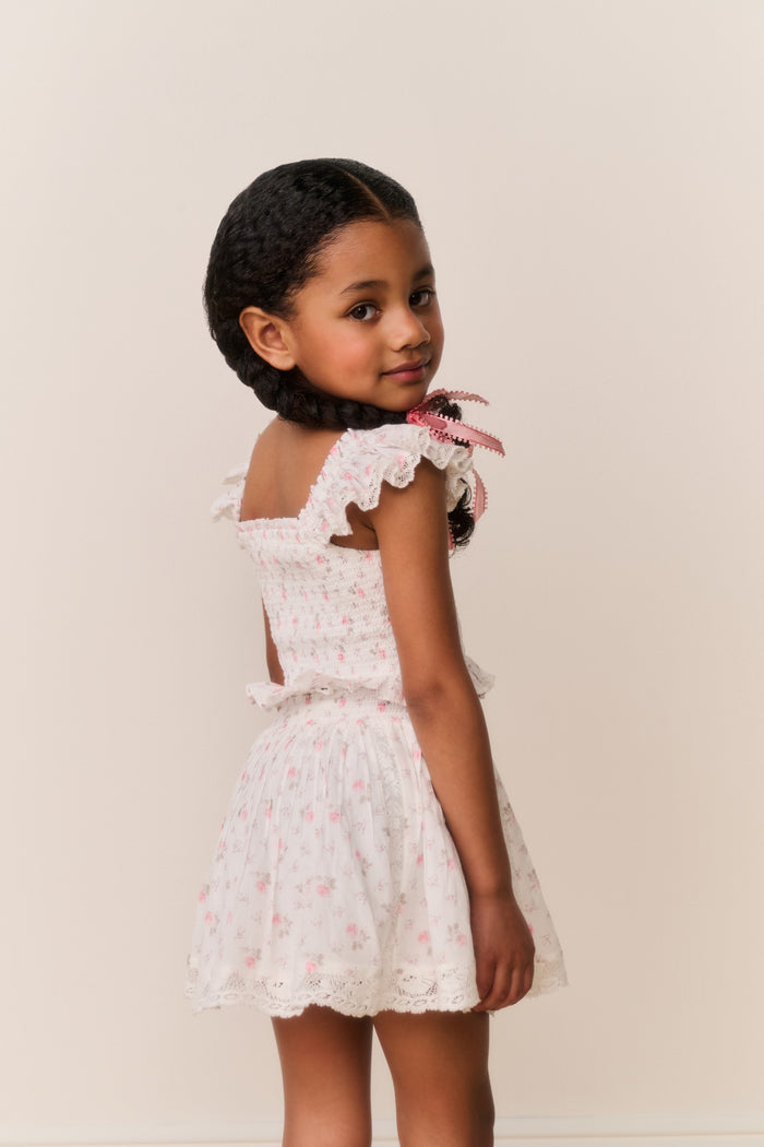 Young girl wearing a white ruffle top and skirt with pink floral patterns on a plain background