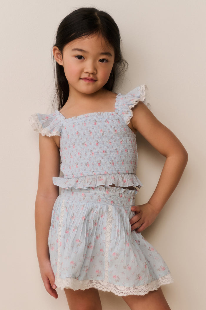Young girl wearing a light blue ruffle hem top and skirt with pink floral patterns on a plain background