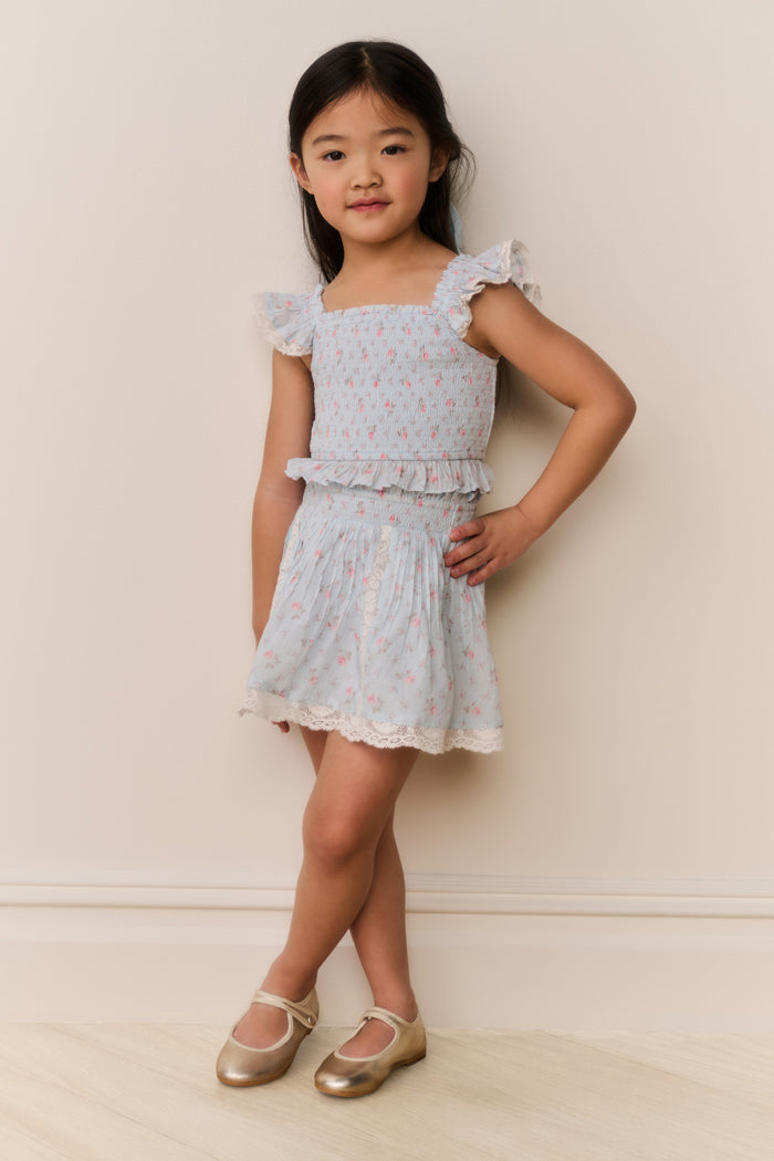 Young girl wearing a light blue ruffle hem top and skirt with pink floral patterns on a plain background