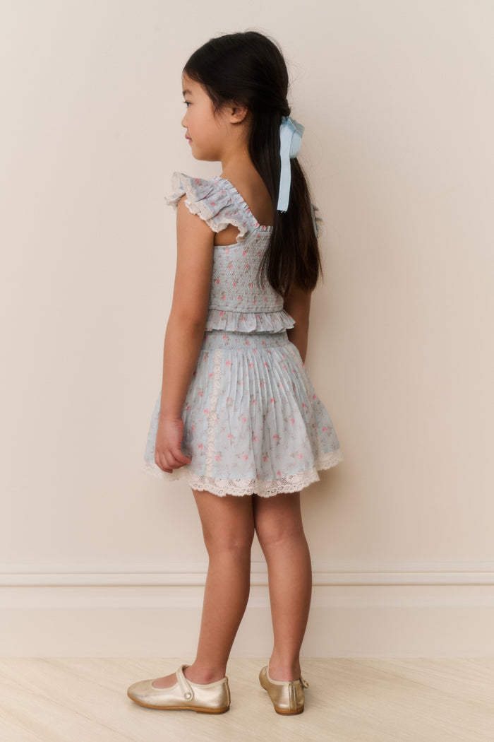 Young girl wearing a light blue ruffle top and skirt with pink floral patterns on a plain background