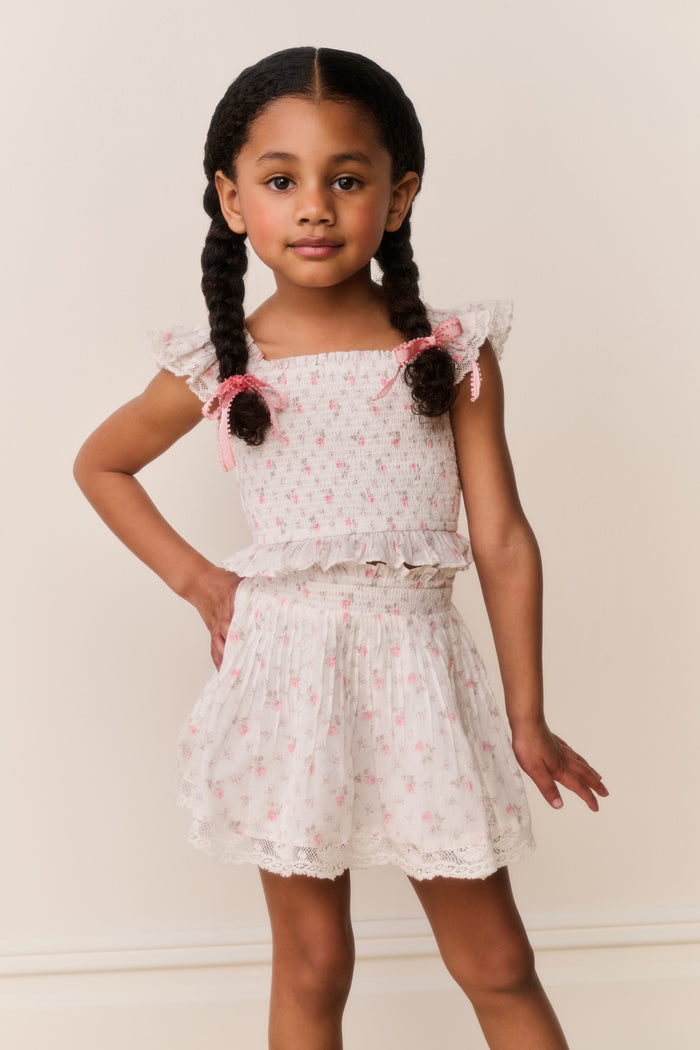 Young girl wearing a white ruffle top and skirt with pink floral patterns on a plain background