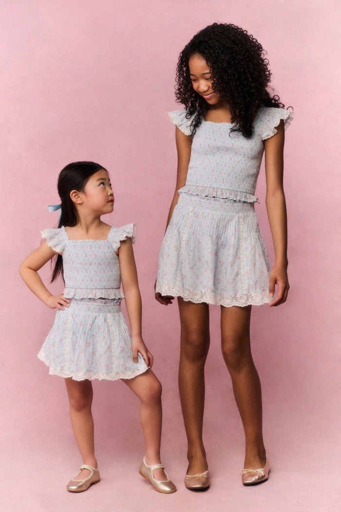 Two young girls wearing matching Light blue floral lace skirt and top against a pink background