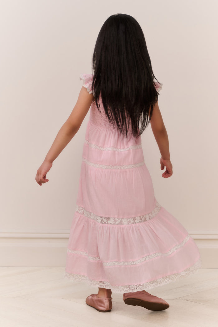 Young girl wearing a pink dress with lace details, standing against a plain background.