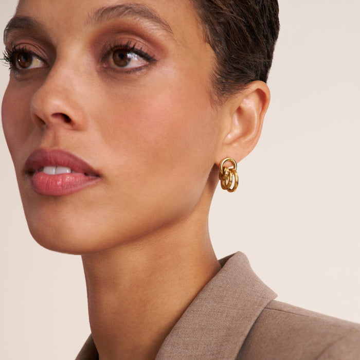 A close-up of a model wearing a gold double-ring hoop earring, shown from the side against a warm beige background. | Lead