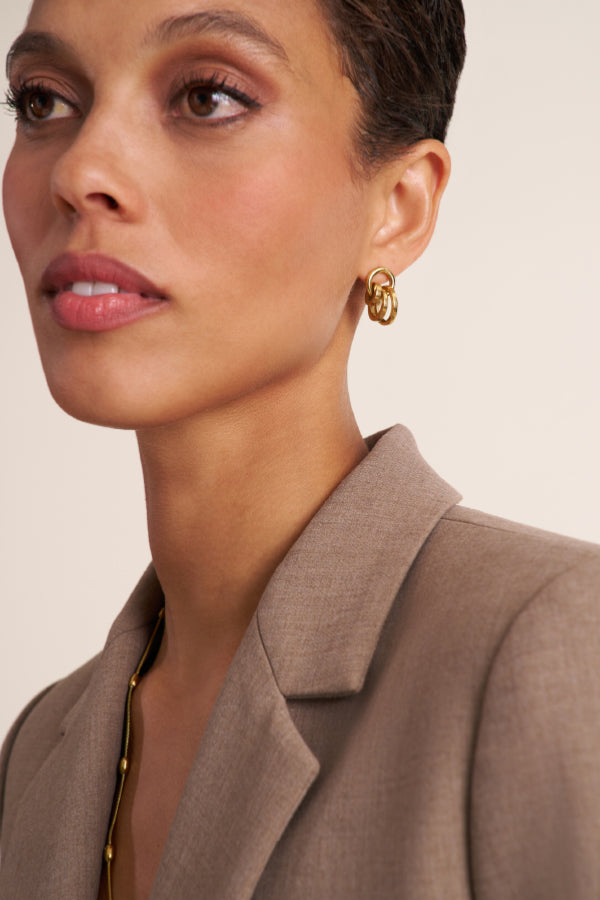 Close-up of a woman wearing a gold geometric hoop earring, styled with a taupe blazer against a warm beige background. | Exclude | Grid Hover