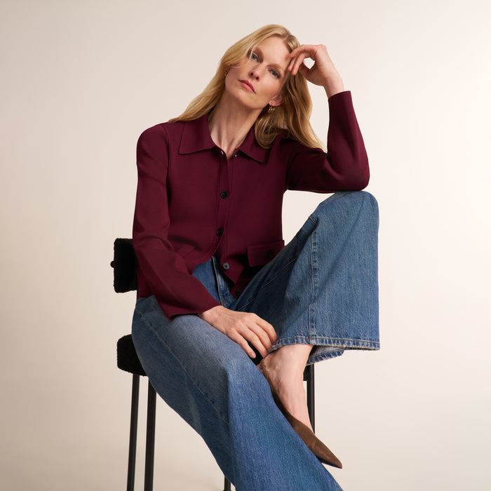 A woman sits on a black stool wearing a long-sleeve burgundy button-up shirt and wide-leg blue jeans against a plain beige studio background. | Lead
