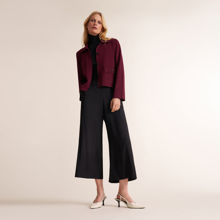 A woman models a dark burgundy cropped jacket over a black turtleneck, paired with black wide-leg cropped trousers and white slingback heels, against a solid light beige studio background.