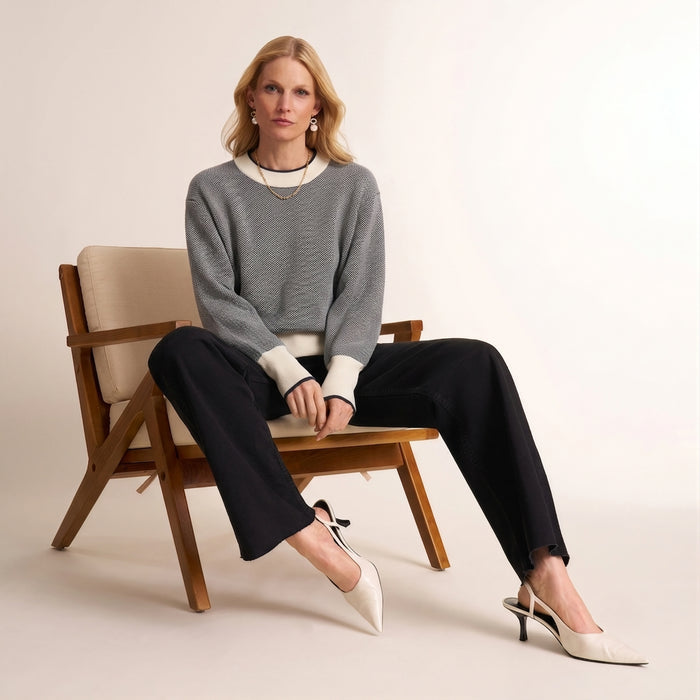 A woman sits on a wooden chair wearing a gray knit sweater with navy blue trim, black wide-leg pants, and white slingback heels against a plain light beige studio background. | Lead