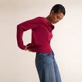 A woman wearing a bright magenta long-sleeve knit cardigan with buttons and blue jeans, posing against a plain beige studio background. | Lead