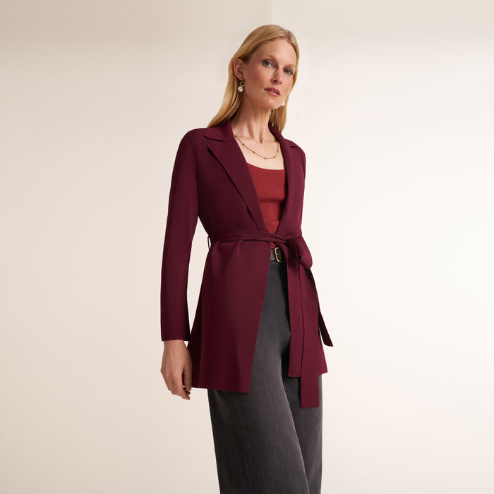 A woman modeling a long-sleeve wrap-style blazer in deep burgundy, worn over a rust-red top and dark gray trousers, against a plain light beige studio background. | Lead