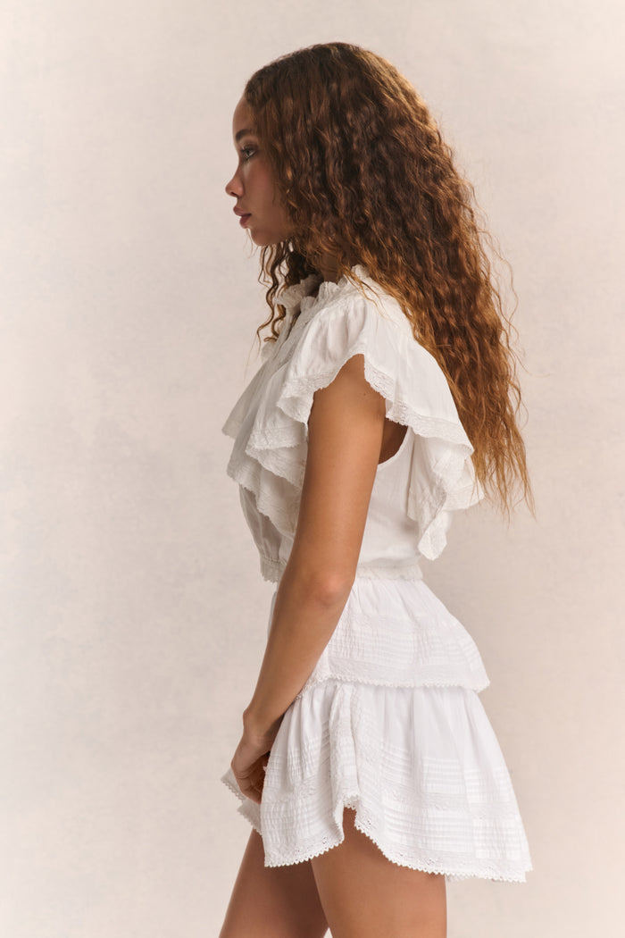 Woman wears white ruffle cropped top with mini white ruffle skirt against beige background