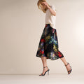 A woman models a black midi skirt with a vibrant, painterly multicolor abstract print, paired with a cream top and black strappy high-heeled sandals, set against a plain beige studio background. | Lead