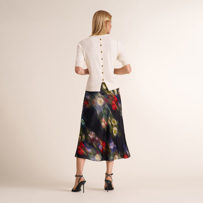 A woman shown from the back wearing a cream short-sleeve jacket with gold buttons and a dark midi skirt with a colorful floral print, paired with black high heels, against a plain beige studio background.