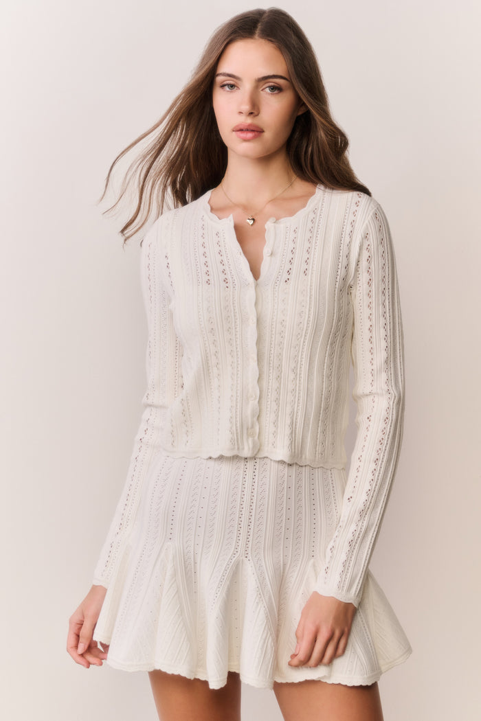Woman wearing a cream-colored cardigan and dress with intricate patterns on a plain background