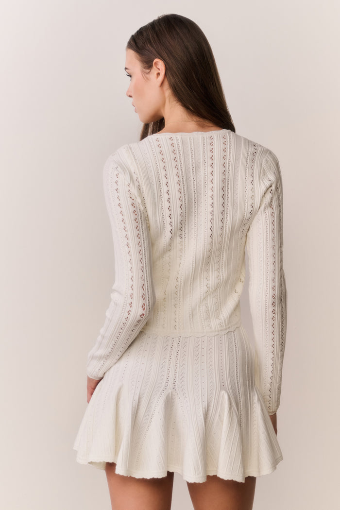 Woman wearing a cream-colored cardigan and dress with intricate patterns on a plain background