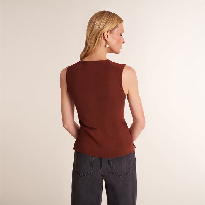 A woman shown from the back wearing a sleeveless rust-brown top and dark gray jeans, standing against a plain light beige studio background.