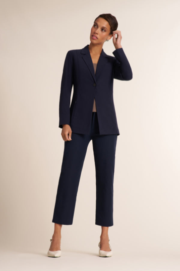 A woman modeling a navy blue two-piece suit consisting of a single-breasted blazer and matching cropped trousers, paired with white pointed-toe heels, against a solid light beige studio background.  | Exclude
