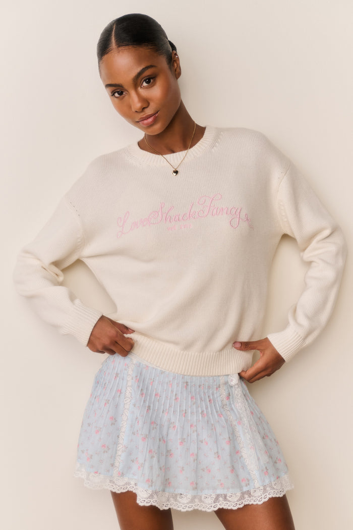 Woman wearing a cream sweater with pink LoveShackFancy text and a blue floral skirt against a beige background