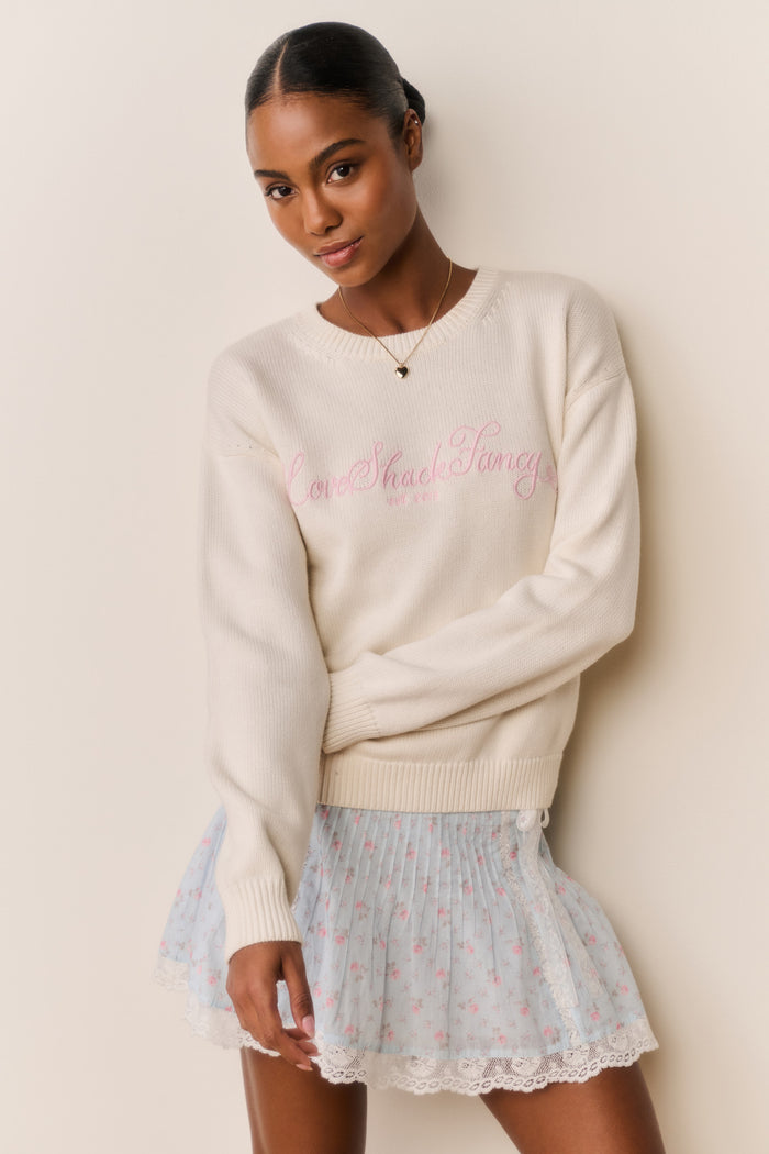 Woman wearing a cream sweater with pink LoveShackFancy text and a blue floral skirt against a beige background